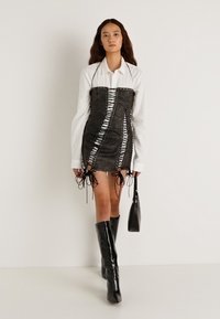 Crna kožna haljina s korsetskim dizajnom, s bijelim vezicama i detaljima vezanja, uparena s bijelom košuljom i crnim čizmama do koljena.