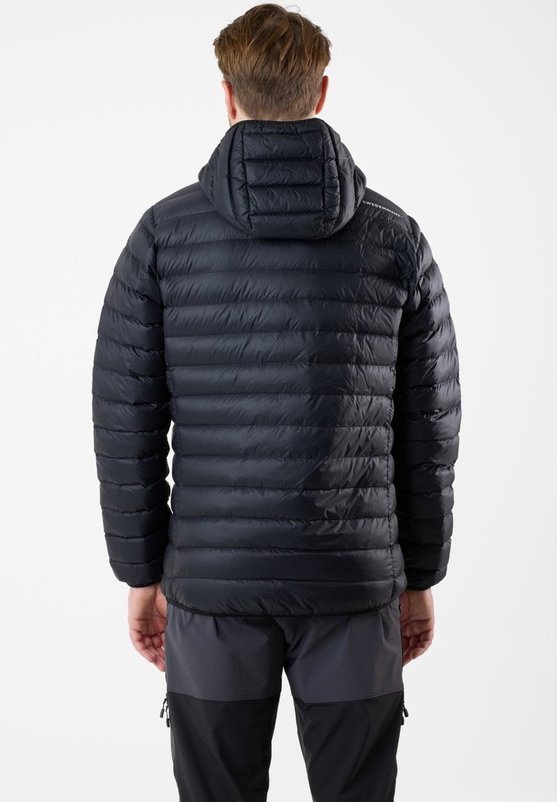 Homme portant une veste matelassée noire à capuche et un pantalon bicolore foncé, vu de dos sur un fond blanc.