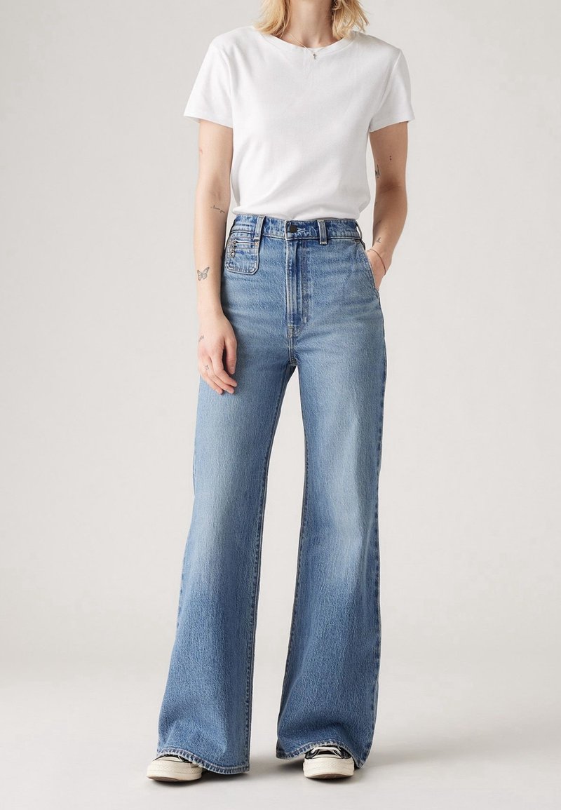 Femme portant un jean bleu clair taille haute à jambes larges, un T-shirt blanc à manches courtes, et des baskets, debout avec une main dans la poche.