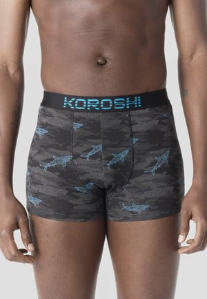 Torso masculino vistiendo bóxers oscuros con estampado de camuflaje y patrones geométricos de tiburones en azul, con una cinturilla negra que lleva la marca "KOROSH" en azul.