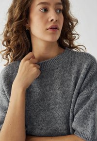 Maglione grigio a maglia con maniche corte e colletto a costine; texture morbida con una vestibilità leggermente ampia; la mano del modello poggia sul colletto.