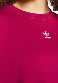 Magenta bluza wykonana z miękkiej bawełny, z małym białym logo Adidas trefoil na górnej lewej stronie. Klasyczny krój z okrągłym dekoltem.