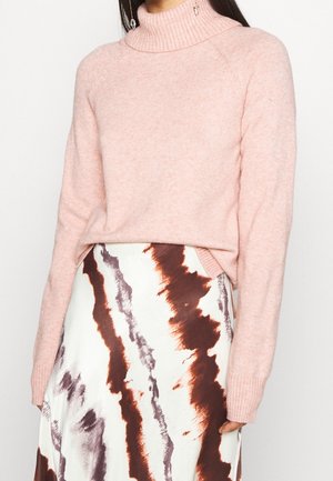 Femme portant un pull à col roulé rose clair associé à une jupe blanche avec des rayures abstraites marron et grises.