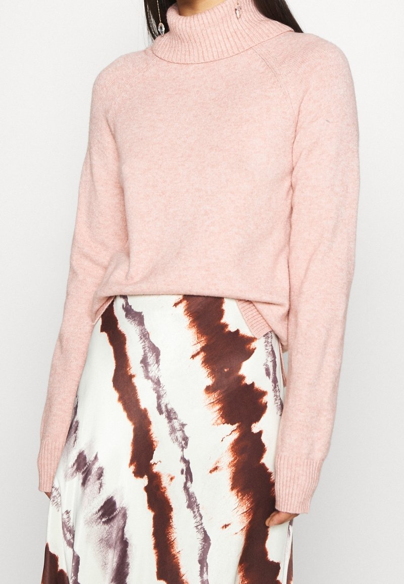 Femme portant un pull à col roulé rose clair associé à une jupe blanche avec des rayures abstraites marron et grises.