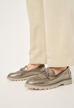 Mocassins en bronze métallique avec un design cousu, un grand accent en chaîne et une semelle texturée et confortable, accompagnés de pantalons beige clair.