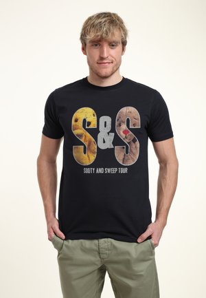 T-shirt in cotone nero con grandi lettere gialle e marroni testurizzate "S&S", accompagnato da un dettaglio grafico di marionette. Il testo sottostante recita "Sooty and Sweep Tour."