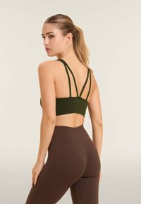 Sujeción deportiva en verde oliva con doble tirante, silueta corta y tejido suave. Combinada con leggings marrones de cintura alta.