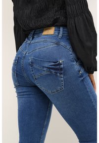 Jeans de denim azul con un diseño ajustado, que cuentan con una cinturilla alta, múltiples bolsillos con costura curva y una etiqueta de marca de cuero en la cintura.