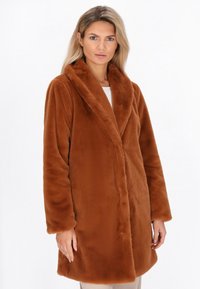Kvinde iført en lang, brun faux fur-frakke med brede klapper, poserende mod en simpel hvid baggrund.