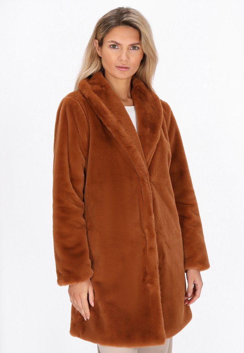 Kvinde iført en lang, brun faux fur-frakke med brede klapper, poserende mod en simpel hvid baggrund.