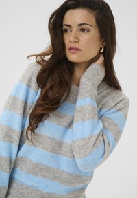 Gaiši pelēks džemperis ar zilām svītrām, apaļu izgriezumu un garām piedurknēm. Mīksta tekstūra, ikdienišķs piegriezums ar ribotiem aprocēm.