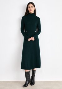 Gestricktes, dunkelgrünes Kleid mit hohem Kragen, langen Ärmeln und silbernen Akzenten an der Taille. Gestylt mit schwarzen, knielangen Stiefeletten mit Absatz.