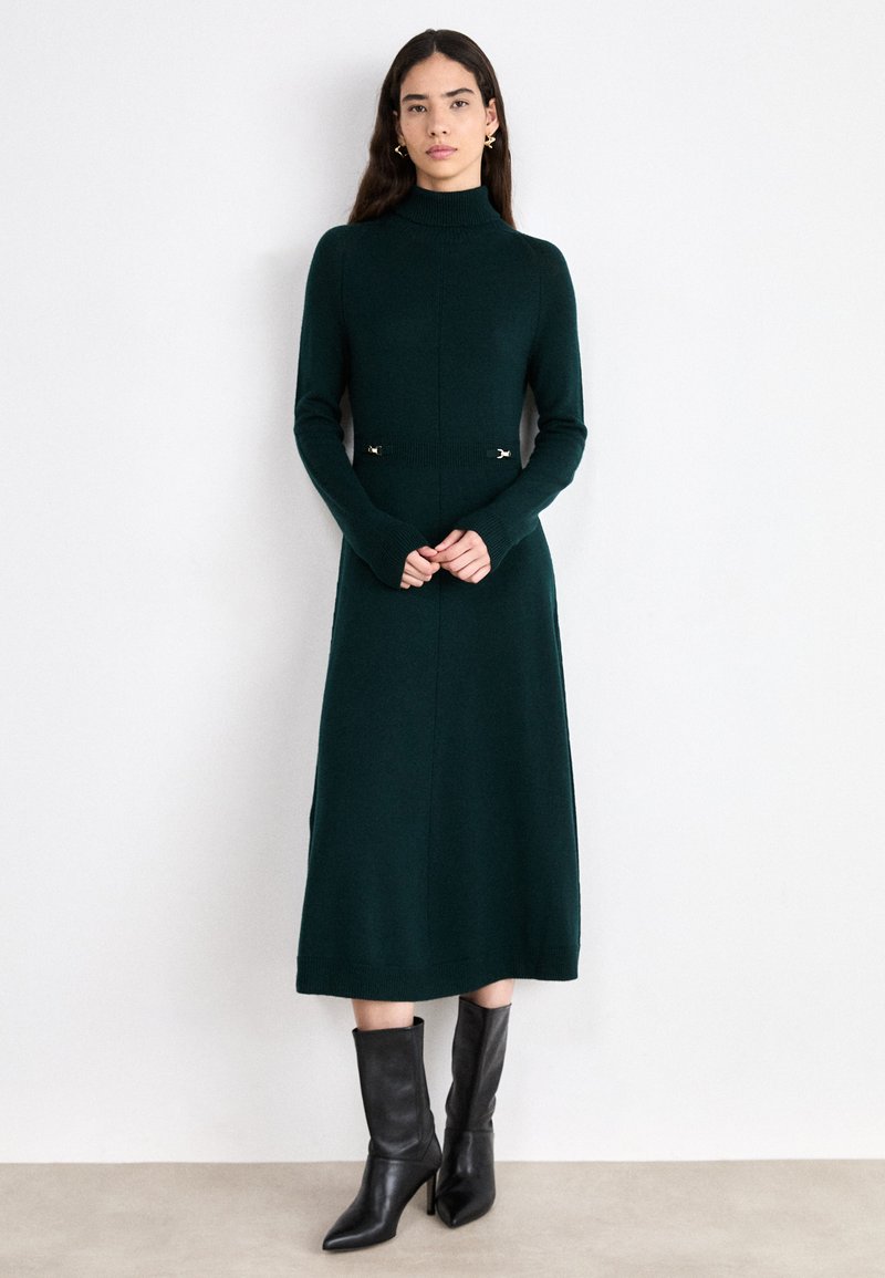 Gestricktes, dunkelgrünes Kleid mit hohem Kragen, langen Ärmeln und silbernen Akzenten an der Taille. Gestylt mit schwarzen, knielangen Stiefeletten mit Absatz.