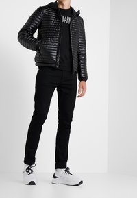 Svart puffjacka med quiltat mönster, huva och dragkedja; kombinerad med svarta jeans och vita sneakers med svarta detaljer.