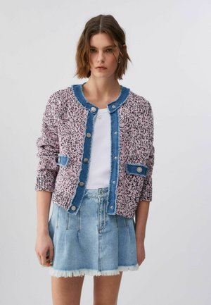 Giovane donna che indossa un cardigan rosa lavorato a maglia con bordi blu e una gonna di jeans azzurro con orlo sfrangiato, su uno sfondo chiaro uniforme.