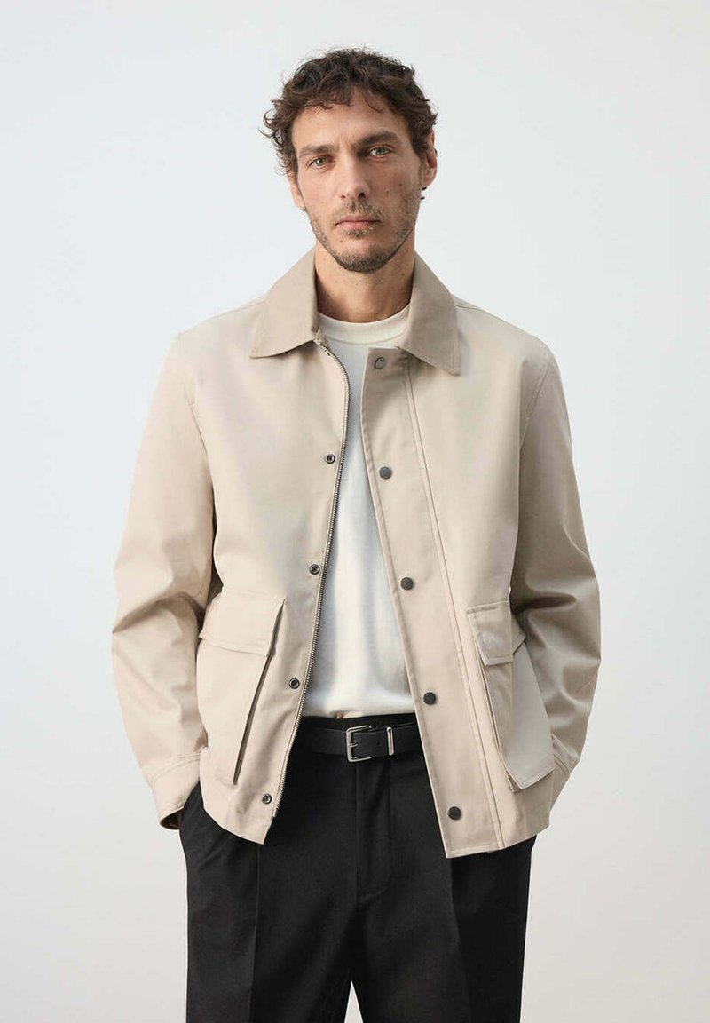Homme aux cheveux courts et bouclés portant une veste beige sur une chemise blanche et un pantalon noir avec une ceinture noire, sur un fond clair uni.