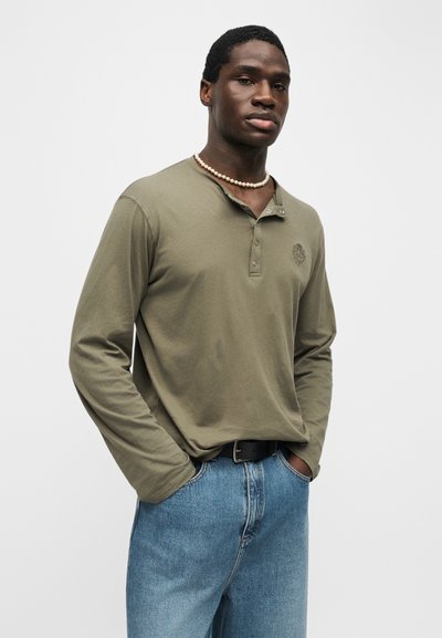 Νεαρός άνδρας ντυμένος με λαδί μακρυμάνικο πουκάμισο henley, μπλε τζιν, μαύρη ζώνη και μαργαριταρένιο κολιέ, όρθιος με τα χέρια στις τσέπες.