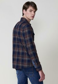 Camicia a quadri in blu navy, rosso e bianco, con colletto a bottone e taschino sul petto. Indossata con jeans in denim blu, caratterizzati da cuciture a contrasto.
