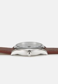 Orologio in metallo argentato con quadrante rotondo e cinturino in pelle marrone. Presenta un quadrante semplice con marcatori delle ore e una corona per la regolazione.