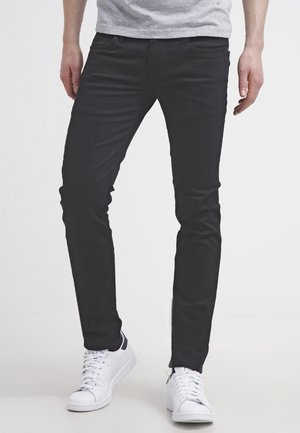 Personne portant un pantalon noir slim et des baskets blanches, debout devant un fond clair uni, les mains détendues le long du corps.