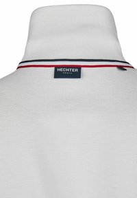 Witte poloshirt van een gestructureerde stof, met een hoge kraag en een label op de achterkant. Rode, zwarte en witte strepen versieren de kraag.