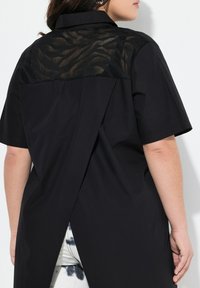 Zwarte shirt met korte mouwen, voorzien van een doorschijnende achterkant met patroon; inclusief een gespleten ontwerp aan de onderkant voor extra detail en textuur.