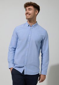 Camicia a maniche lunghe di colore blu chiaro, realizzata in un tessuto morbido, con un logo ricamato discreto sul petto e un colletto classico.