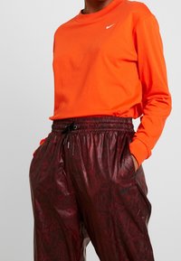 Långärmad orange Nike-tröja kombinera med mörkröda byxor i ormskinnsmönster, med resårmidja och snördetaljer.