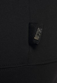 Tessuto nero con texture a coste, con un piccolo etichetta in tessuto nell'angolo con un logo metallico. Design semplice e elegante.