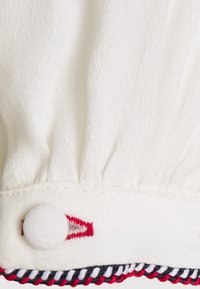 Tessuto bianco con texture arricciata, dotato di chiusura a bottone rotondo e un bordo decorativo di cuciture rosse e blu navy.