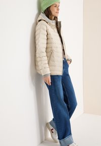 Doudoune beige avec capuche et doublure grise, portée sur un haut clair et un jean large bleu, associée à des baskets blanches avec des accents rouges.