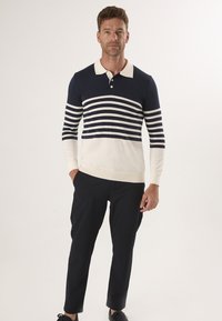 Polo w paski w kolorze granatowym i kremowym, z długimi rękawami i kołnierzykiem, z guzikami. Noszony z ciemnymi spodniami i casualowymi butami. Gładka dzianinowa faktura.