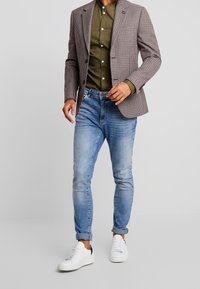 Man i rutigt kavaj, olivgrön skjorta med knappar, ljusblå uppvikt jeans och vita sneakers som står mot en grå bakgrund.