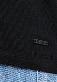 Material negru cu o mică etichetă dreptunghiulară cusută pe care scrie „BIG STAR,” deasupra blugilor din denim albastru, la marginea inferioară.