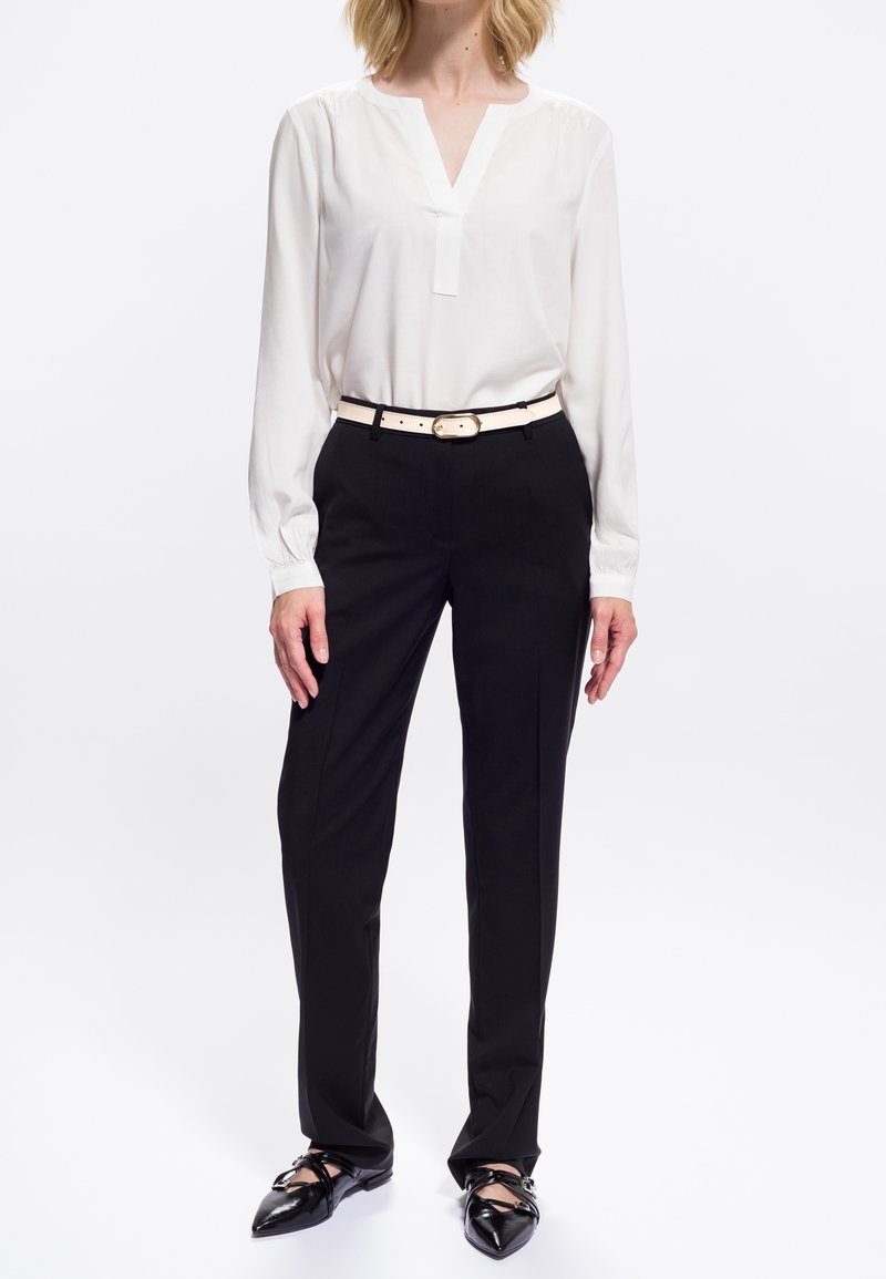 Femme portant un chemisier blanc à manches longues, un pantalon noir droit, une ceinture blanche et des ballerines noires à bout pointu avec des brides.