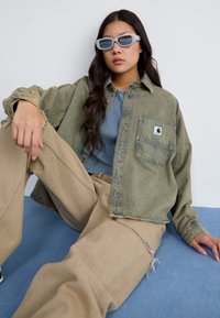 Denim shirt in faded green, featuring a front pocket, paired with beige pants. The model wears light blue sunglasses and a blue top.