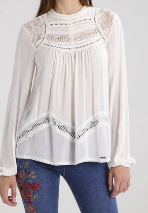 Biała bluzka z długimi rękawami, ozdobiona koronkowymi detalami na ramionach i dole, wykonana z teksturowanej tkaniny, o luźnym kroju.