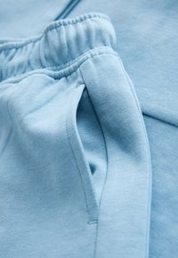 Vaaleansininen kangas, jossa on pehmeä rakenne. Varustettu joustavalla vyötärönauhalla ja sivusauman taskulla. Yksinkertainen muotoilu ilman näkyviä kuvioita tai koristeita.