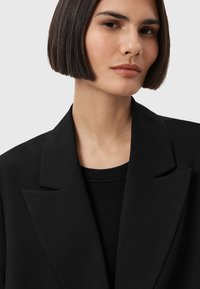 Blazer nero con rever affilati e una maglia nera testurizzata sotto. Tessuto liscio, design strutturato e silhouette moderna.