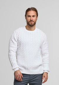 Witte kabelgebreide trui gemaakt van gestructureerde stof, met een ronde halslijn en geribde manchetten en zoom, gedragen over donkere broek.