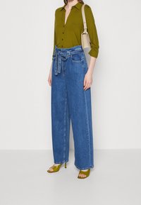 Olivenfarvet skjorte med knaplukning, højtaljede viddejeans i blå med et bundet bælte, og grønne hæle-sandaler. Enkel og afslappet påklædning.