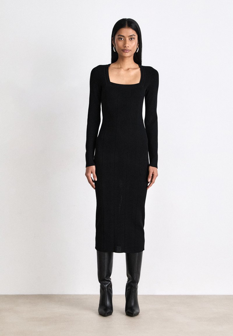 Long-sleeve black knit dress with a square neckline, textured pattern, and fitted silhouette, paired with sleek black ankle boots.