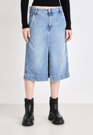 Jupe midi en denim bleu clair avec fente frontale, portée avec un haut noir à manches longues et des bottines noires sur un fond uni.