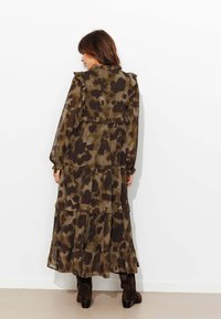 Robe longue à volants, à motifs marron et verts, en tissu transparent, avec des épaules volantées et des manches longues, associée à des bottes hautes marron foncé.