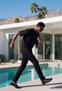 Uomo con abbigliamento nero e occhiali da sole che cammina accanto a una piscina all'aperto con palme e montagne sullo sfondo.