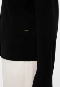 Maglione nero con una morbida texture a maglia, caratterizzato da un'etichetta con logo dorato sull'orlo e polsini a coste. Sullo sfondo si vedono pantaloni di colore chiaro.