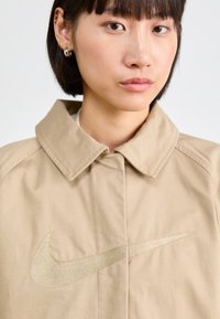 Casaco bege com colarinho clássico e um logo Nike bordado em bege na parte da frente. Textura de tecido suave com um design estruturado.