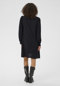 Robe noire longueur genou avec manches longues bouffantes et coupe ample. Tissu avec une texture subtile. Associée à des bottes noires jusqu'au genou.