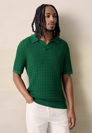 Mann mit langen geflochtenen Haaren, der ein strukturiertes grünes Polohemd mit kurzen Ärmeln und weiße Hose trägt, steht mit einer Hand in der Tasche.