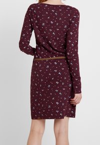 Langärmliges Kleid in tiefem Burgunderrot mit einem floralem Muster in Blau und Weiß. Es hat eine figurbetonte Silhouette und einen geflochtenen Gürtel in der Taille.